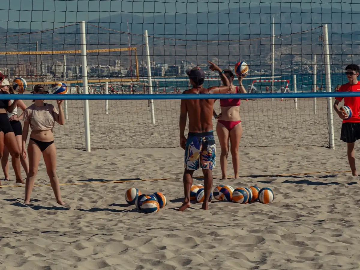 Grupo de jóvenes entrenando voley playa junto a la red, con balones en la arena