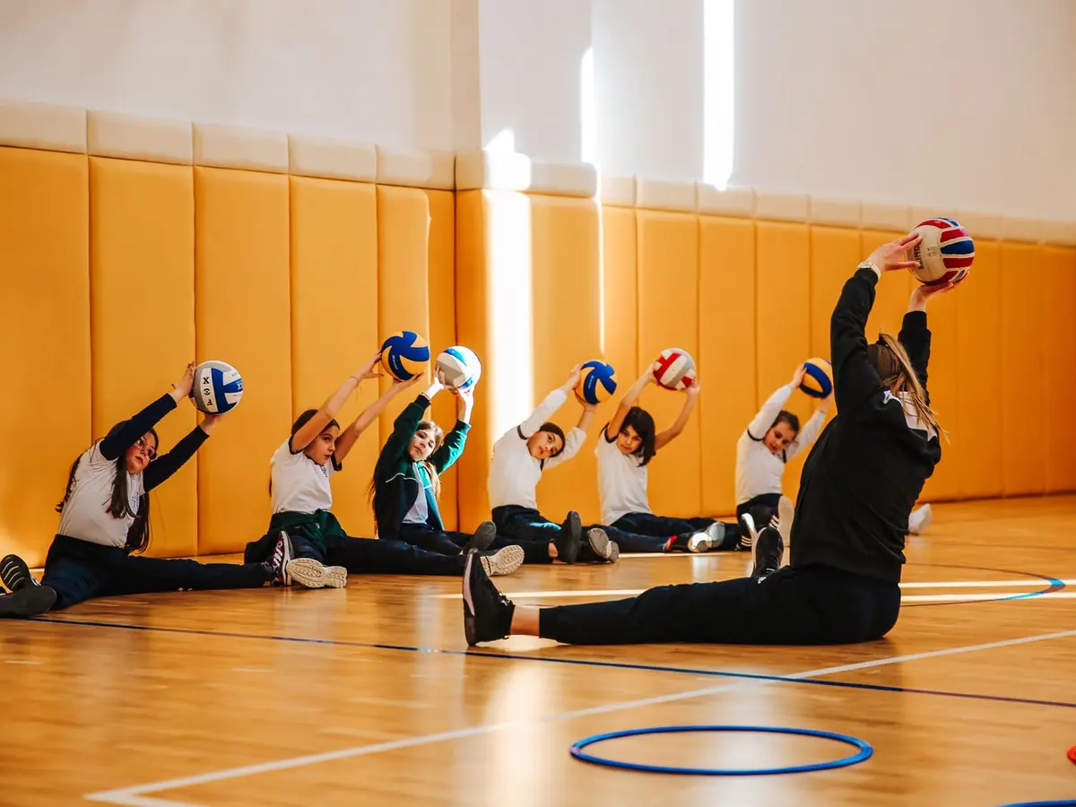 School pupils stretching with volleyballs in a sports hall, led by their coach